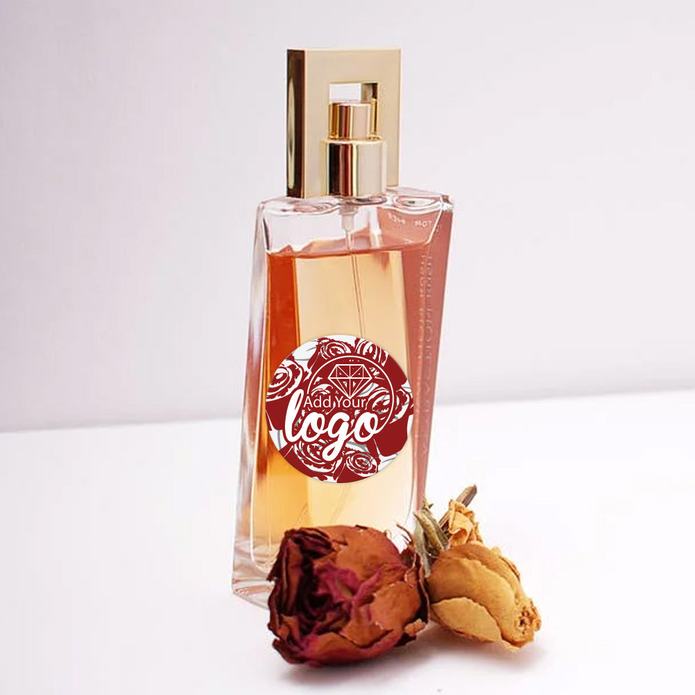 A flower themed label is affixed on a perfume bottle. There are delicate flowers in front of the perfume. The grey background is contrasting against the labeled perfume label.